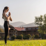 Yogalehrerin Sandra Kohns praktiziert den Baum (Vrksasana) auf einer grünen Wiese bei blauem Morgenhimmel – Symbol für Prana im Yoga und innere Balance im Leben.
