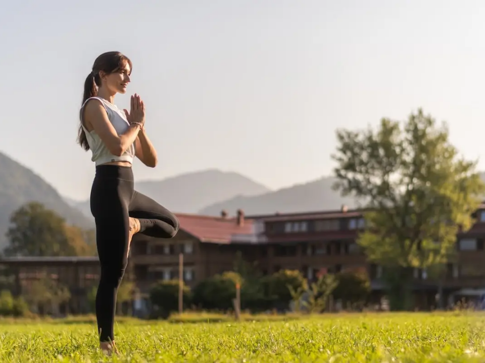 Yogalehrerin Sandra Kohns praktiziert den Baum (Vrksasana) auf einer grünen Wiese bei blauem Morgenhimmel – Symbol für Prana im Yoga und innere Balance im Leben.