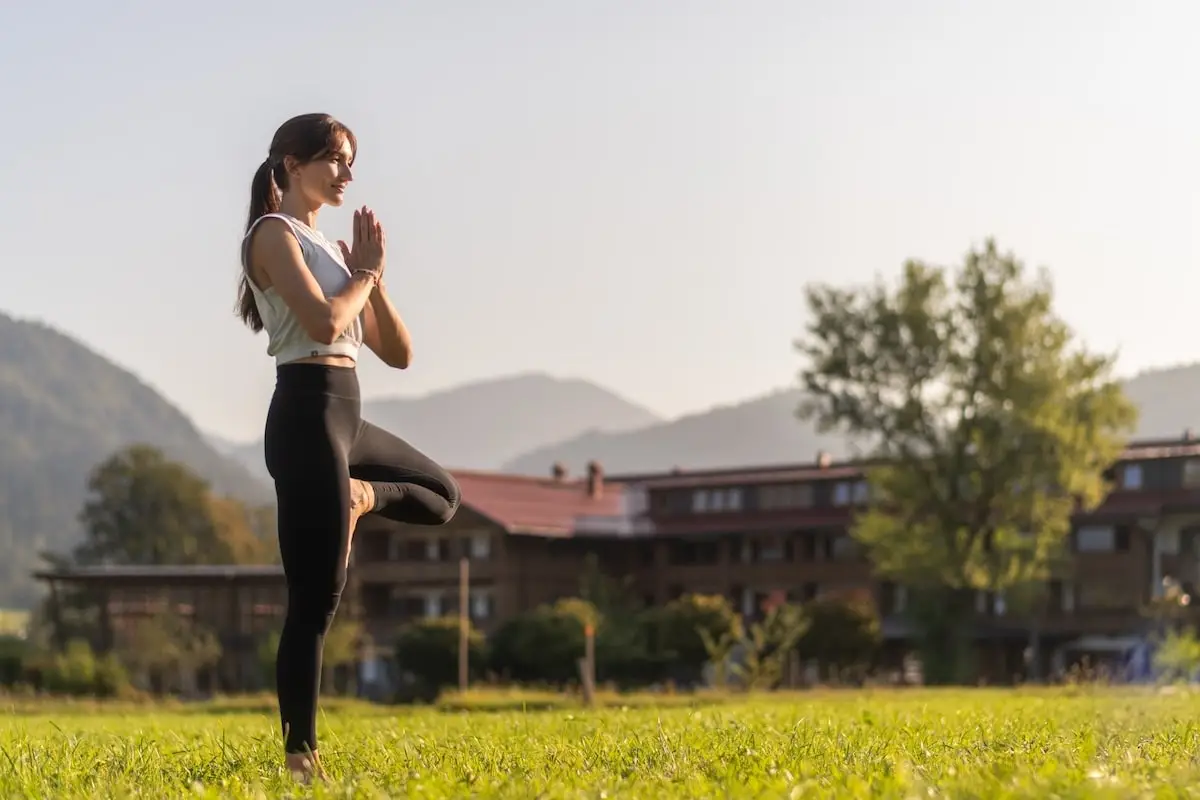 Yogalehrerin Sandra Kohns praktiziert den Baum (Vrksasana) auf einer grünen Wiese bei blauem Morgenhimmel – Symbol für Prana im Yoga und innere Balance im Leben.